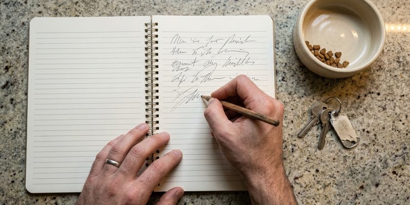 Una mano escribe con lápiz en una libreta junto a un cuenco y unas llaves